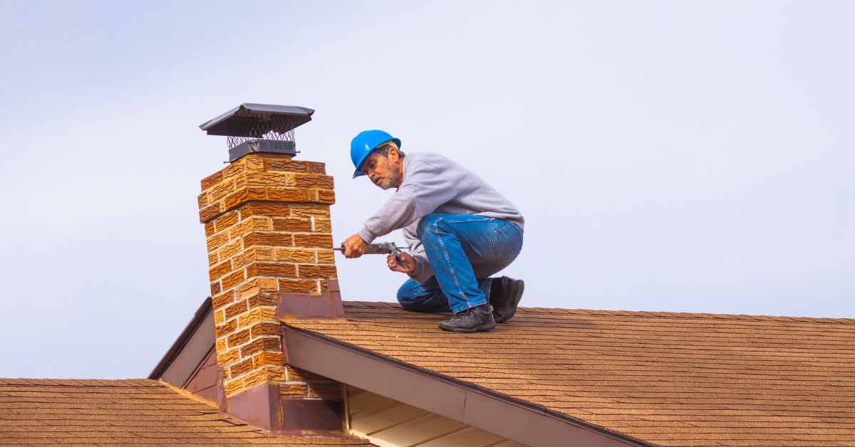 man-repairing-chimney-on-roof-how-to-know-when-its-time-for-chimney-repair-den-defenders