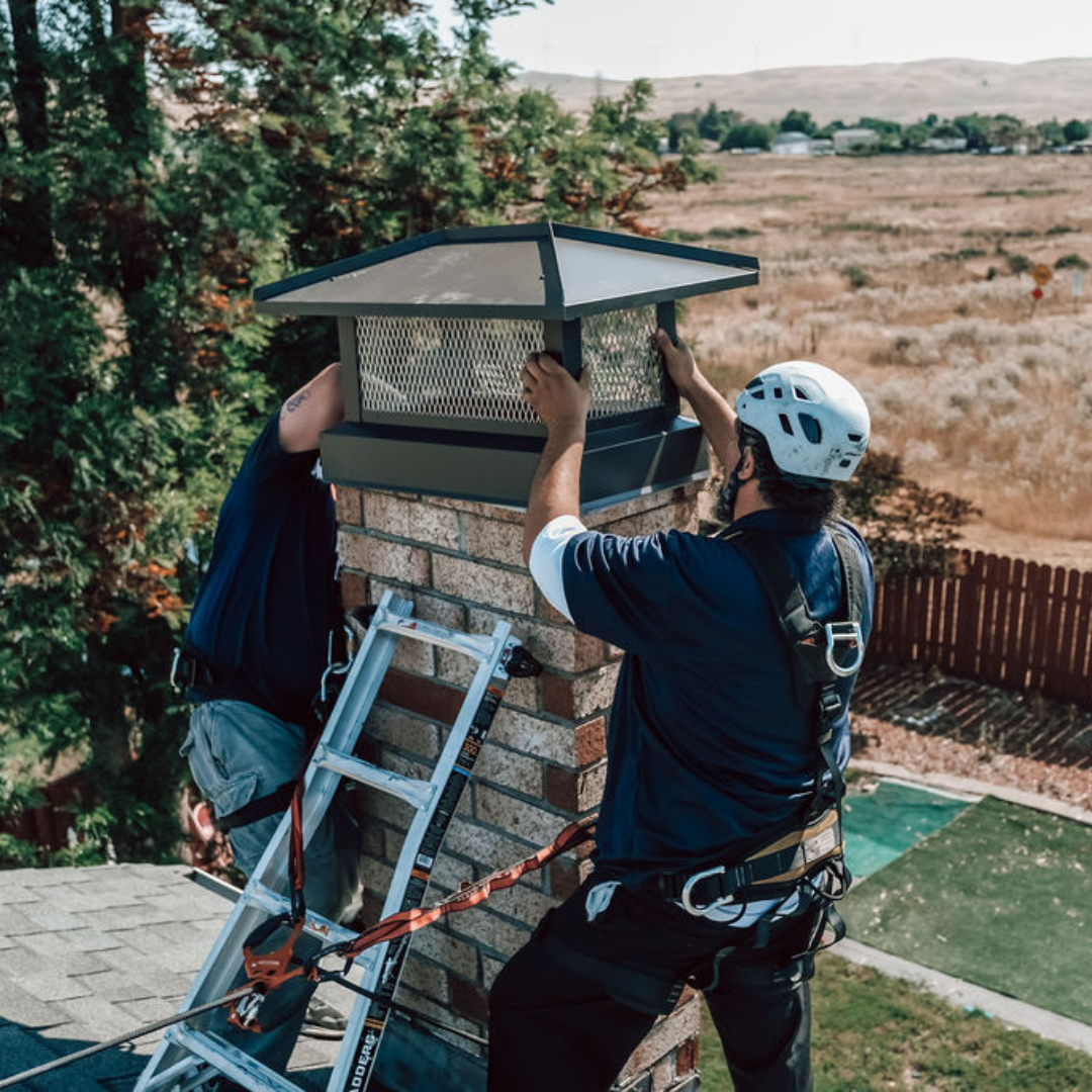 Two Den Defenders Chimney & Fireplace technicians installing a metal chimney cap on a masonry chimney
